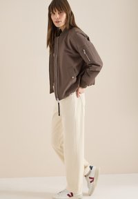 Femme portant une veste aviateur marron, un pantalon large crème et des baskets blanches avec des accents rouges et marines, debout devant un fond beige.
