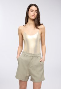 Body in tessuto metallico oro senza maniche abbinato a pantaloni corti a vita alta di un verde oliva chiaro, dotati di tasche laterali e una texture liscia.