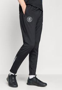 Sorte treningsbukser laget av glatt stoff med en avsmalnende design, sidelommer med glidelås og en sirkulær FC-logo på venstre lår.