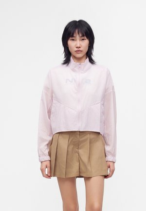 Jeune femme aux cheveux noirs portant une veste zippée rose clair et un short plissé beige sur un fond blanc uni.