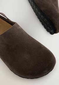 Braune Wildlederschuhe mit offenem Rücken, flacher schwarzer Gummisohle, glatter Textur und runder Zehenform. Verfügt über einen kleinen Absatz für Stabilität.