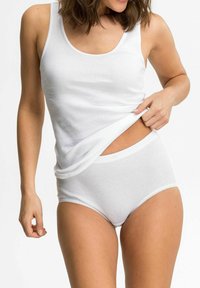 Woman wearing white cotton tank top and high-waisted underwear lifting hem to reveal waist against a plain background.