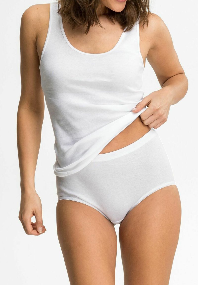 Woman wearing white cotton tank top and high-waisted underwear lifting hem to reveal waist against a plain background.