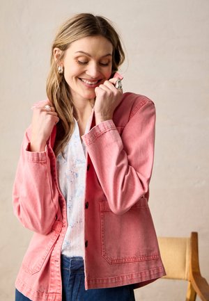 Smiling woman wearing a pink buttoned jacket over a white patterned blouse and blue jeans, holding jacket collar with both hands.