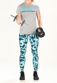 Camiseta deportiva gris con texto azul, leggins turquesa con estampado y zapatillas negras, sosteniendo mancuernas en cada mano.