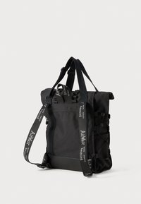 Zaino nero realizzato in tessuto resistente, dotato di spallacci regolabili, chiusura a rotolo, tasche laterali e una cinghia decorativa con testo.