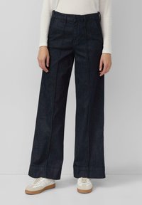 Weite dunkelblaue Jeanshose mit vertikalen Nähten, hoher Taille und umgeschlagenen Säumen. Kombiniert mit weißen Sneakers.