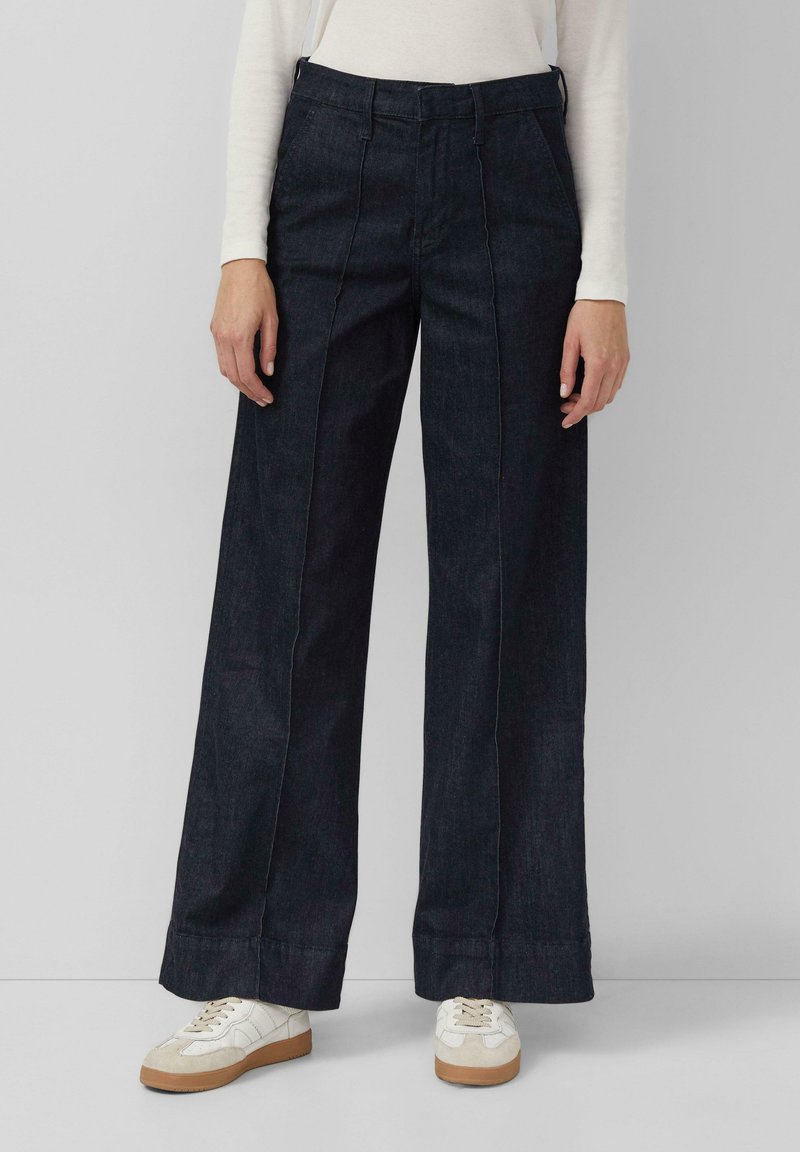 Weite dunkelblaue Jeanshose mit vertikalen Nähten, hoher Taille und umgeschlagenen Säumen. Kombiniert mit weißen Sneakers.