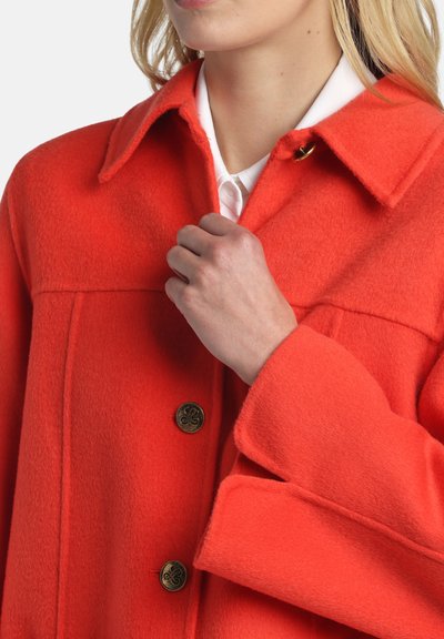 Žena v jasnom oranžovom vlnenom kabáte s veľkými gombíkmi, drží golier blízko bielej košele pod ním.