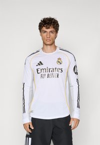 Hvid langærmet trøje med gule detaljer, sorte striber på ærmerne, har "Emirates Fly Better" logo og Real Madrid våbenskjold.