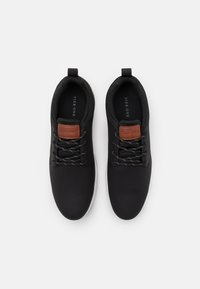 Zwarte sportschoenen met een glad bovenmateriaal, een bruin leren accent op de tong en grijze veters boven een witte zool.