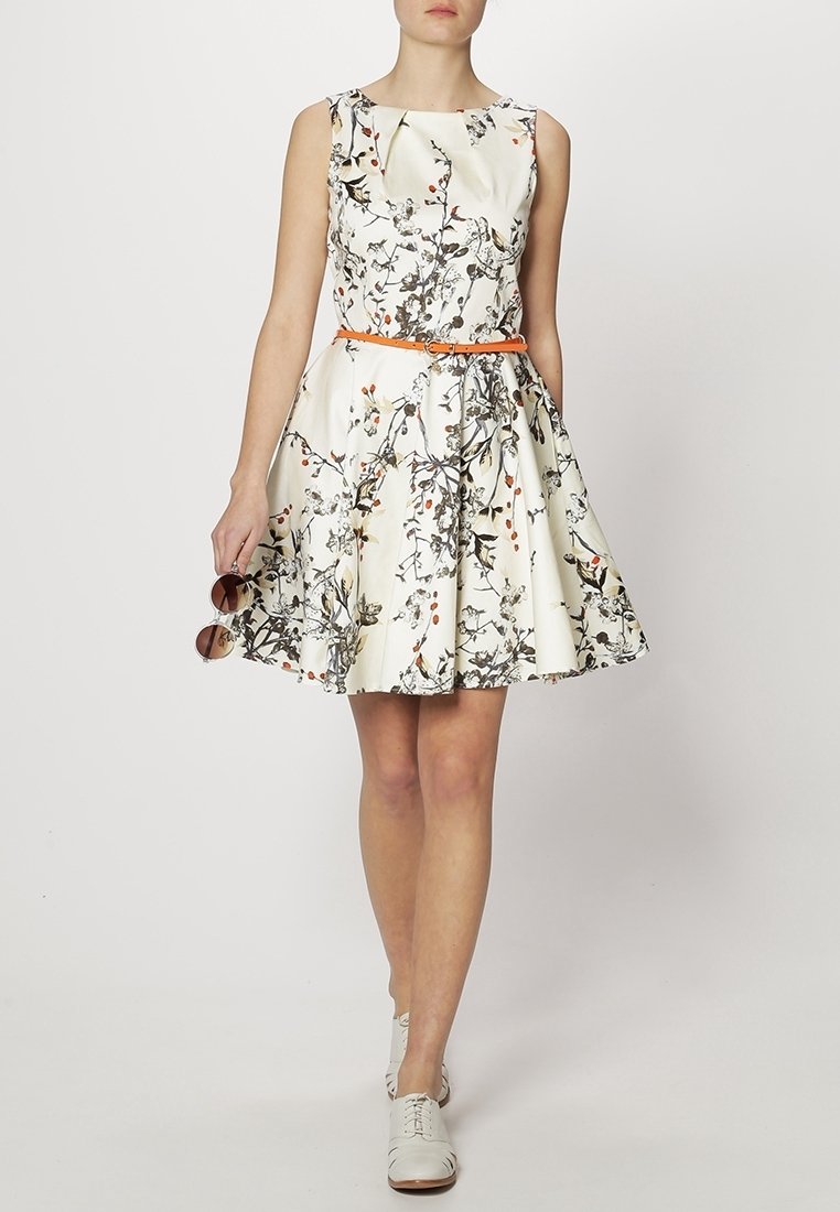 Femme portant une robe blanche sans manches à motifs floraux avec une ceinture orange, tenant des lunettes de soleil, et des chaussures blanches à lacets, se tenant devant un fond uni.
