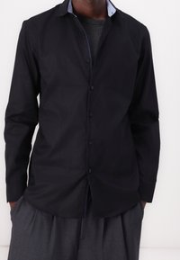 Camicia nera a maniche lunghe con chiusura frontale a bottoni, caratterizzata da un colletto azzurro chiaro con motivo e una texture liscia. Indossata con pantaloni grigi.