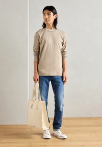 Bež majica s long sleeves in okroglim izrezom, v kombinaciji z modrimi jeans hlačami. Drži smetano bež platneno torbo, nosi bele čevlje in stoji na lesenem tleh.
