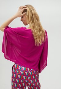 Haut sheer magenta avec de larges manches. Associé à un pantalon multicolore à motif géométrique avec des accents rouges, bleus et crème.