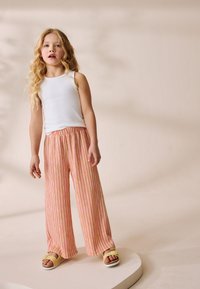 Débardeur blanc, pantalon pattes d'éléphant rayé rose et jaune, sandales jaunes. Tissu lisse avec une coupe décontractée. Fond clair.