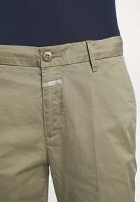 Kaki bomullschinos med en mjuk textur, som har en framficka, knappstängning och en liten märkesetikett nära fickan.