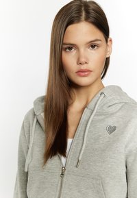 Šedá mikina na zip s designem srdce na hrudi. Obsahuje kapuci s šňůrkou a přední kapsy. Hladká textura látky.