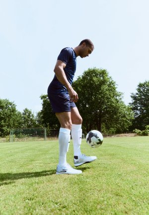 Joueur de football masculin en tenue bleu marine et chaussettes blanches jongle avec le ballon sur un terrain herbeux avec des arbres et un but en arrière-plan.