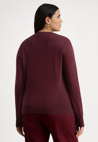 Γυναίκα με μακριά σκούρα μαλλιά που φοράει χρυσούς κρίκους στα αυτιά, πουλόβερ μακρυμάνικο μπέργκουντι και παντελόνι μπέργκουντι, στραμμένη προς τα πλάγια, με λευκό φόντο.