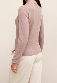 Vrouw in een ribbelige, mauvekleurige trui met lange mouwen en hoge taille witte broek, gezien van achteren en van opzij.