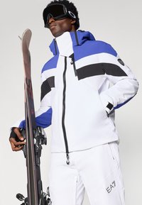 Valge suusajakk siniste ja mustade detailidega, millel on kõrge krae, eesmine tõmblukk ja logo paan varrukal. Käes on lume suusad.