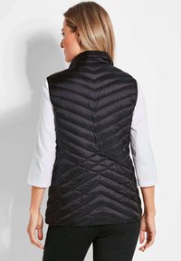 Gepolsterte schwarze Weste mit hohem Kragen und Zick-Zack-Steppmuster, von hinten betrachtet, kombiniert mit einem weißen Langarmhemd.