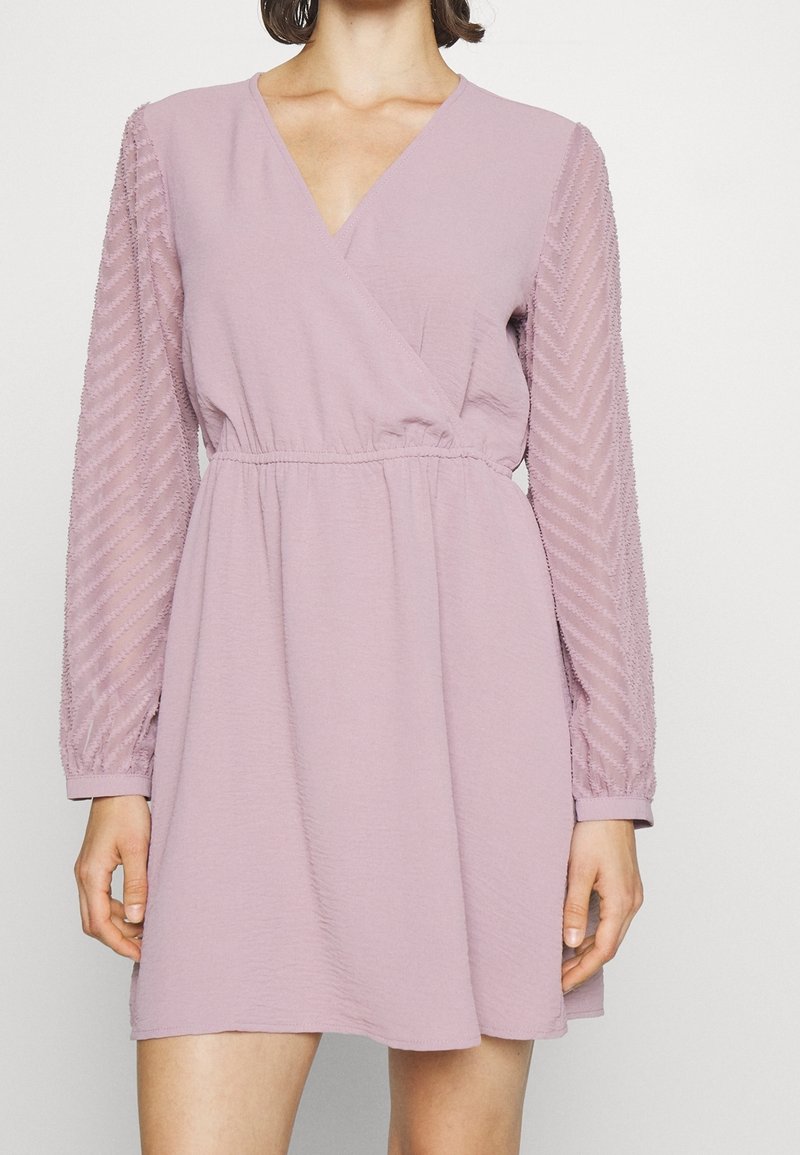 Robe portefeuille lilas clair avec un décolleté en V profond, des manches longues en voile transparent avec un motif en chevrons, et une taille ajustée pour mettre en valeur la silhouette.
