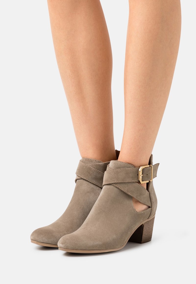Bottes en daim taupe montant à la cheville avec talons carrés et sangles enveloppantes maintenues par des boucles dorées, portées sur des jambes nues sur un fond clair.