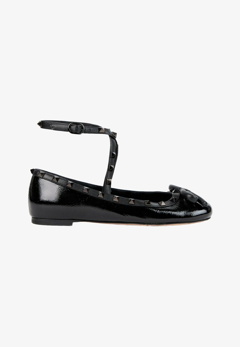 Chaussure plate en cuir verni noir avec bride à la cheville, clous pyramidaux noirs le long des bords et un nœud sur l’orteil.