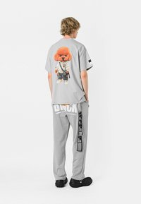 Camiseta gris claro con un perro de dibujos animados vestido con un traje blanco en la parte de atrás. Combinada con pantalones grises a juego que tienen texto y un detalle de correa.