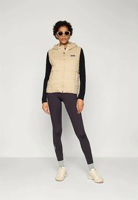 Vesta puffer beige cu glugă și închidere cu fermoar, purtată peste o cămașă neagră cu mânecă lungă și colanți negri. Încălțări ușoare și ochelari de soare supradimensionați.