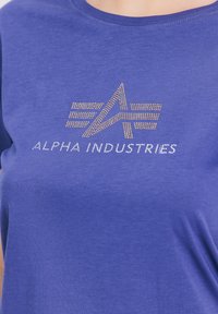 Vijolična majica, izdelana iz mehkega blaga, s krožnim izrezom, ki ima teksturiran zlato-srebrn logotip z napisom "ALPHA INDUSTRIES."