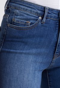 Jeans de mezclilla azul de media altura con una textura suave, que cuentan con un pequeño bolsillo frontal, costuras en color cobre y herrajes metálicos en la cinturilla.