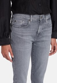 Jeans in denim grigio con un design aderente, cinque tasche e leggeri segni di usura. Abbinati a una blusa nera con dettagli in pizzo.
