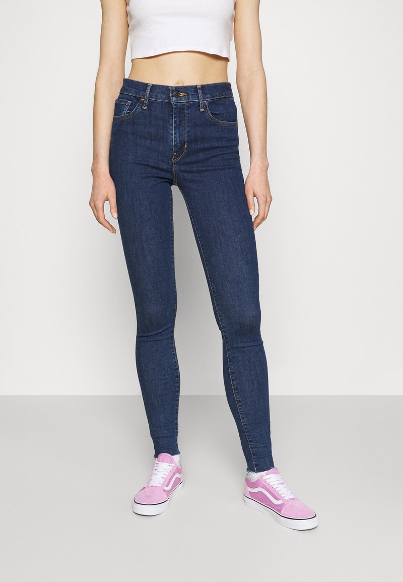 Personne portant un jean skinny taille haute bleu foncé, un top court blanc et des baskets roses et blanches, debout devant un fond uni.