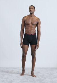 Melni bokseršorti ar sarkanu "HUGO" jostas joslu, pieguļošs dizains. Modelis stāv neitrālā pozā uz mīksta, gaiši pelēka virsmas.