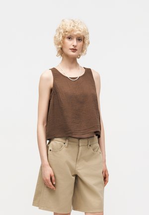 Ξανθό άτομο που φοράει αμάνικη καφέ υφασμάτινη μπλούζα και μπεζ σορτς μέχρι το γόνατο, στέκεται μπροστά σε απλό ανοιχτό φόντο.