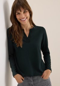 Haut long à manches longues de couleur vert foncé avec un col mandarin et une texture douce, associé à un pantalon gris. Le mannequin se tient devant un mur clair.