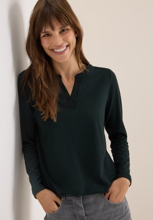 Haut long à manches longues de couleur vert foncé avec un col mandarin et une texture douce, associé à un pantalon gris. Le mannequin se tient devant un mur clair.