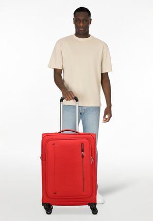 Red rolling suitcase with a textured surface, a front pocket, and a retractable handle. It features four wheels for easy maneuverability.
