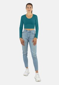 Türkisen langärmligen Cropped-Top, kombiniert mit hellblauen High-Waist-Jeans und weißen Sneakers, präsentiert an einem weiblichen Modell.