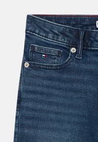 Temno modri kavbojki iz džins materiala z sprednjim žepom, na katerem je majhna oznaka v rdeči, beli in modri barvi, s srebrnim gumbom za zapiranje ter diskretnim bledenjem.