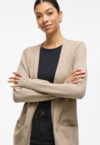 Beige strikket cardigan med åpen front, ribbede mansjetter og to frontlommer, parret med en svart topp og mørkeblå jeans. Glatt tekstur.