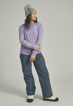 Frau trägt eine graue Mütze, ein lavendelfarbenes Langarmshirt mit weißen Grafiken, dunkelblaue Hose und weiße Slip-On-Schuhe, steht mit verschränkten Armen.