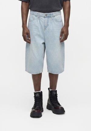 Person trägt hellblaue kniehohe Jeansshorts, schwarze hohe Schnürstiefel und ein dunkles Hemd mit entspannt an den Seiten hängenden Armen.