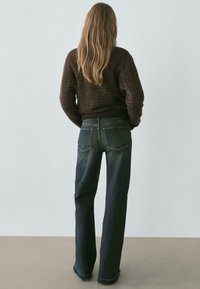 Kobieta o długich włosach stoi twarzą do gładkiej ściany, ubrana w ciemnobrązowy sweter i wyblakłe niebieskie dżinsy o prostych nogawkach.