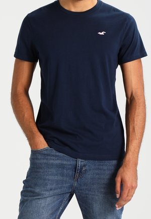 Mand iført navyblå kortærmet t-shirt med lille hvidt logo på brystet og blå jeans, med den ene hånd i lommen, foran ensfarvet baggrund.