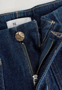 Tumši zili džinsi no denim auduma ar misiņa rāvējslēdzēju, metāla pogu aizdari un redzamām šuvju detaļām. Iekšējā etiķete daļēji redzama.