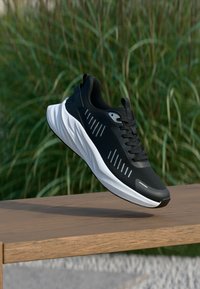 Schwarz-weißer Sportschuh schwebt über einer Holzbank vor grünem Laubhintergrund und hebt das Schuhdesign sowie Details der Sohle hervor.